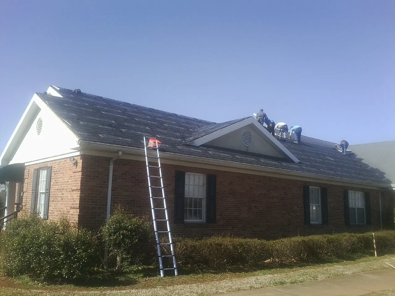 Crew tearing shingles off a brick ranch roof with a ladder set — Metal Roof Repair work by our crew in Batavia